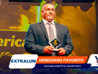 Smiling man in a dark suit on stage, holding a glass trophy, with a bright, colorful backdrop and event banners behind him.