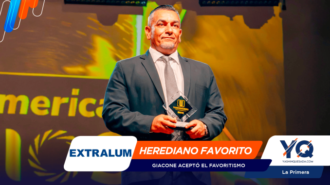 Smiling man in a dark suit on stage, holding a glass trophy, with a bright, colorful backdrop and event banners behind him.