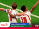 Two soccer teammates celebrate with arms around each other on a green field as fans or players celebrate nearby (informative moment).