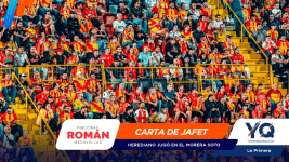Crowded stadium stands with fans wearing red and yellow jerseys, yellow railings and stairways visible in the background, foreground shows sponsor banners.