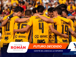 Soccer players in yellow jerseys form a huddle with arms around each other on a stadium field with a crowd in the stands behind them