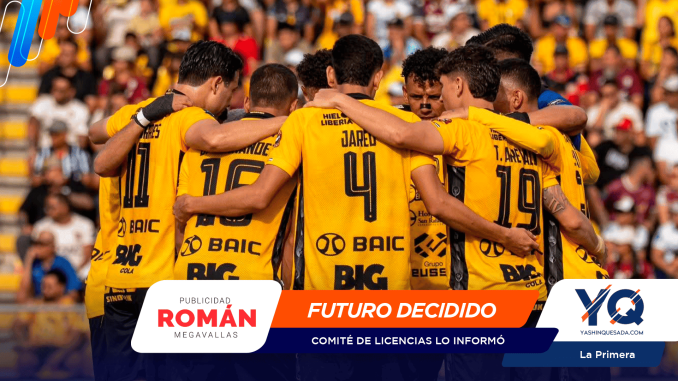 Soccer players in yellow jerseys form a huddle with arms around each other on a stadium field with a crowd in the stands behind them