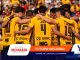 Soccer players in yellow jerseys form a huddle with arms around each other on a stadium field with a crowd in the stands behind them