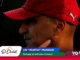 Close-up profile of a man wearing a red cap with a white Puma logo against a dark background with colorful shapes in the corner.