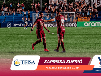 Two soccer players in maroon uniforms celebrate on a green pitch while a cheering crowd watches from blue grandstands behind them.