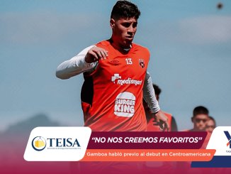 Soccer player in a red jersey with number 13 mid-action on the field against a blue sky, advertising banners blurred at the bottom.