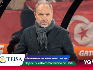 Man in a dark coat and gray scarf seated on a television sports set with red banners and sponsor logos in the background