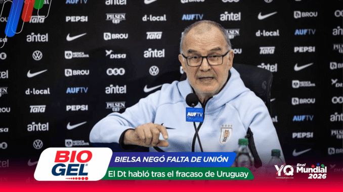 Older man in a light blue jacket speaks into a microphone labeled AUFTV at a press conference, with a sponsor-filled backdrop behind him.