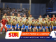 Soccer team in yellow-blue patterned uniforms posing for a group photo on the field with spectators in the stands behind them.