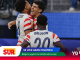 Belgian players celebrate on the court, Balogun #20 embraced by teammates amid blue advertising backdrop.