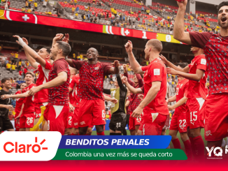 Soccer players in red uniforms celebrate on the field, raising their arms as fans cheer in the stadium stands behind them.