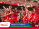 Soccer players in red uniforms celebrate on the field, raising their arms as fans cheer in the stadium stands behind them.