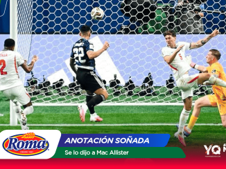 Football match action near the goal: players contesting the ball as it heads toward the net, with a goalkeeper in orange making a save; sponsor banner with Roma logo at bottom.