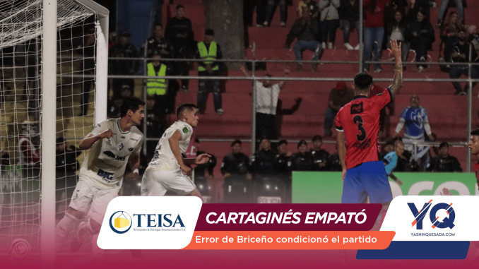 Soccer match scene near the goal: a player in red with number 3 raises his arm while two players in white move toward the goal, with spectators in the stands behind a barrier.