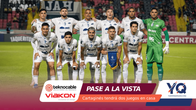 Soccer team posing for a group photo on the field; white uniforms, goalkeeper in green, stadium stands in background.