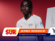 Soccer player in a white RB Leipzig jersey stands against a red studio backdrop with bold overlapping sponsor graphics in orange, blue, and white.