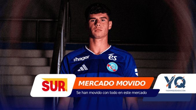 Portrait of a young man in a blue soccer jersey standing in a dim tunnel, sponsor banners partly covering the bottom of the frame.
