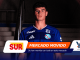 Portrait of a young man in a blue soccer jersey standing in a dim tunnel, sponsor banners partly covering the bottom of the frame.