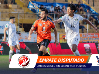 Two soccer players chase the ball during a match in a stadium; one in orange kit, one in white, with spectators and ads in the background,