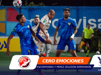 Soccer players in blue and white uniforms compete for a ball in the air on a bright indoor field, with advertising banners in the background.