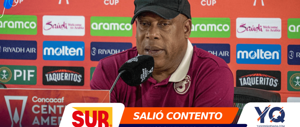Man in a burgundy polo and black cap speaking into a microphone at a press conference with sponsor banners behind him.