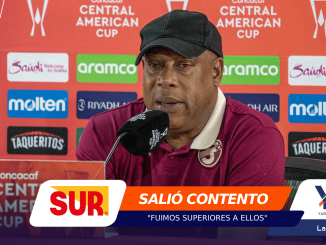 Man in a burgundy polo and black cap speaking into a microphone at a press conference with sponsor banners behind him.