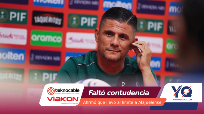 Man in a green shirt at a post-match press conference, touching his ear, with a colorful sponsor backdrop behind him.