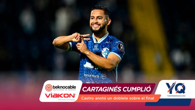 Soccer player in a blue jersey smiling and gesturing on the field, with sponsor banners at the bottom.