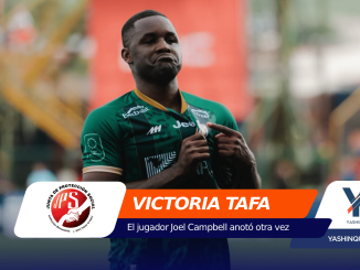 Soccer player in a green jersey adjusting his shirt on a blurred outdoor field, with orange and blue sponsor banners in the foreground reading 'VICTORIA TAFA'.