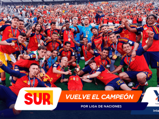 Soccer team in red jerseys posing with medals and a trophy after winning, as cheering fans fill the stands behind them.
