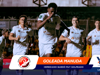 Soccer players in white jerseys celebrate a goal on the field under stadium lights, with banners and branding at the bottom.