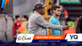 Coach in a gray blazer points to the right on the sideline, with event banners and logos across the bottom of the scene.