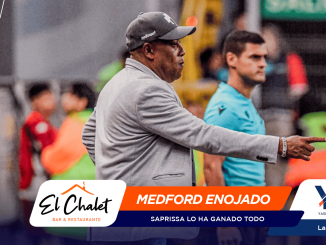Coach in a gray blazer points to the right on the sideline, with event banners and logos across the bottom of the scene.
