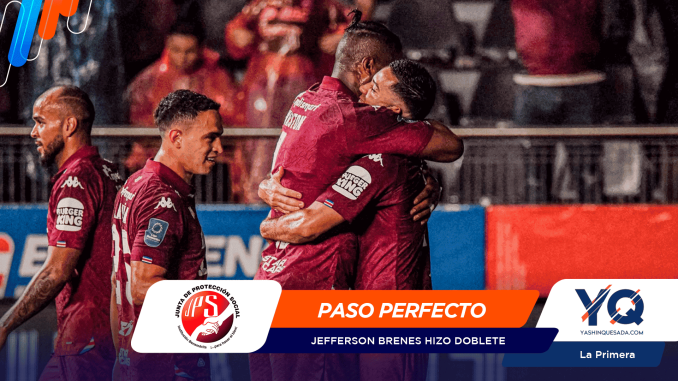 Soccer players in maroon uniforms celebrate on the field, embracing and smiling after a goal.