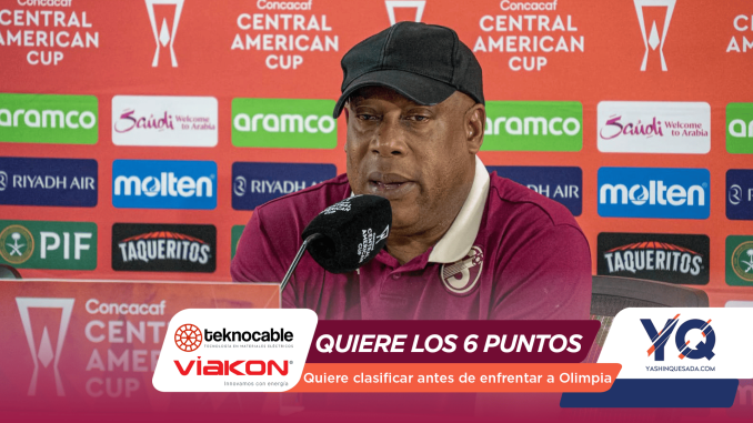Man in a dark cap and maroon polo speaks into a microphone at a press conference, with a red sponsor-filled backdrop behind him.