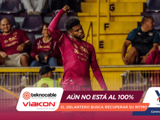 Soccer player in a magenta kit raising a fist in celebration on the pitch, with yellow barriers and seated fans in the background.