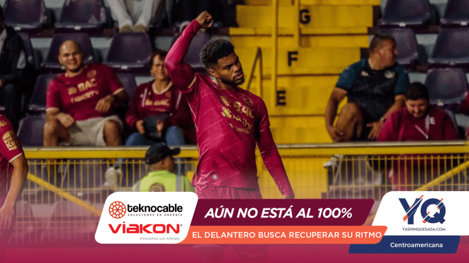 Soccer player in a magenta kit raising a fist in celebration on the pitch, with yellow barriers and seated fans in the background.