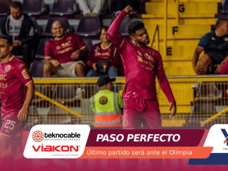 Two soccer players in maroon jerseys on the field, one raising his arm as spectators watch from the stands behind him.